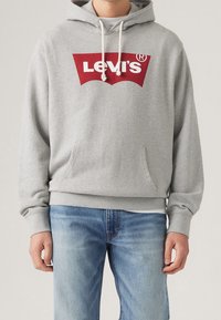 Sweat à capuche gris avec un grand logo Levi's rouge sur le devant. Comprend une poche kangourou et des cordons blancs sur la capuche. Associé à un jean bleu.