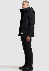 Must hüppeline must puffer-jakk, millel on kapuuts, tõmblukuga rinna tasku ja elastsed varrukad. Kandmise all tumedad teksad ja mustad nööpidega saapad, millel on logotäht.