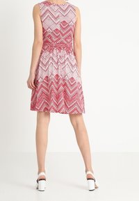 Robe sans manches rouge et blanche avec un motif en zigzag, taille cintrée et jupe évasée ; portée avec des sandales à talons blancs.