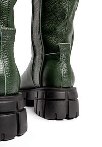 Bottes à cheville en cuir texturé vert foncé avec semelle noire épaisse et languettes arrière, sur fond blanc.