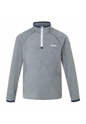 Lächelnder Junge, der eine graue Fleecejacke mit Reißverschluss, marineblaue Hose und schwarze sowie neongrüne Wanderschuhe trägt, steht vor einem schlichten Hintergrund.