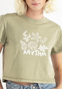 Camiseta recortada de color verde oliva hecha de material suave, que presenta un estampado gráfico blanco de conchas y corales con la palabra "RITMO" debajo.
