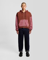 Sudadera con cremallera marrón con acentos de tela de cuadros, que cuenta con un bolsillo frontal y capucha. Combinada con pantalones de mezclilla oscuros con puños enrollados.