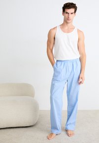 Hellblaue, karierte Schlafanzughosen mit einem elastischen Bund, kombiniert mit einem weißen, ärmellosen Baumwolltanktop. Das bekleidete Model steht barfuß.