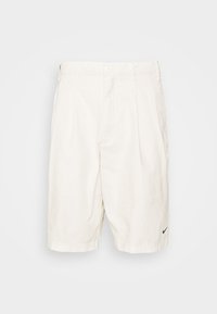 Světlé béžové bavlněné šortky s plisovaným přední částí, dvěma postranními kapsami a malým černým logem Nike na dolní levé straně.