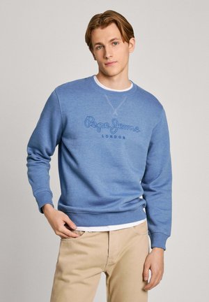 Sudadera azul de tejido suave con cuello redondo, que presenta un logotipo bordado. Lleva puesta sobre una camisa blanca y se combina con pantalones de color beige claro.