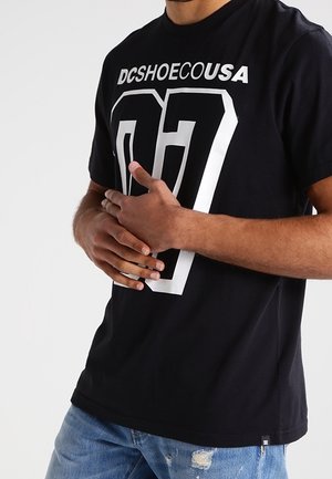 Sort DC Shoes t-shirt med stort hvidt "07" nummer, båret af en person med korslagte hænder, kombineret med revnede blå jeans på en ensartet baggrund.