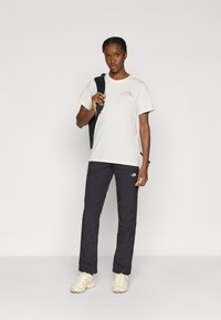 Μπεζ βαμβακερό T-shirt με μικρό λογότυπο, συνδυασμένο με μαύρο νάιλον παντελόνι με λογότυπο, και μπεζ αθλητικά παπούτσια.