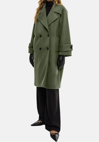 Grøn uldblanding dobbeltbrystet frakke med store klapper og sorte knapper, parret med sorte bukser og quiltede sorte handsker.