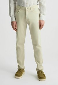 Jeans de algodón color beige claro con un corte ajustado, bolsillos sutiles y cierre de un solo botón, combinados con zapatos verde oliva con cordones.