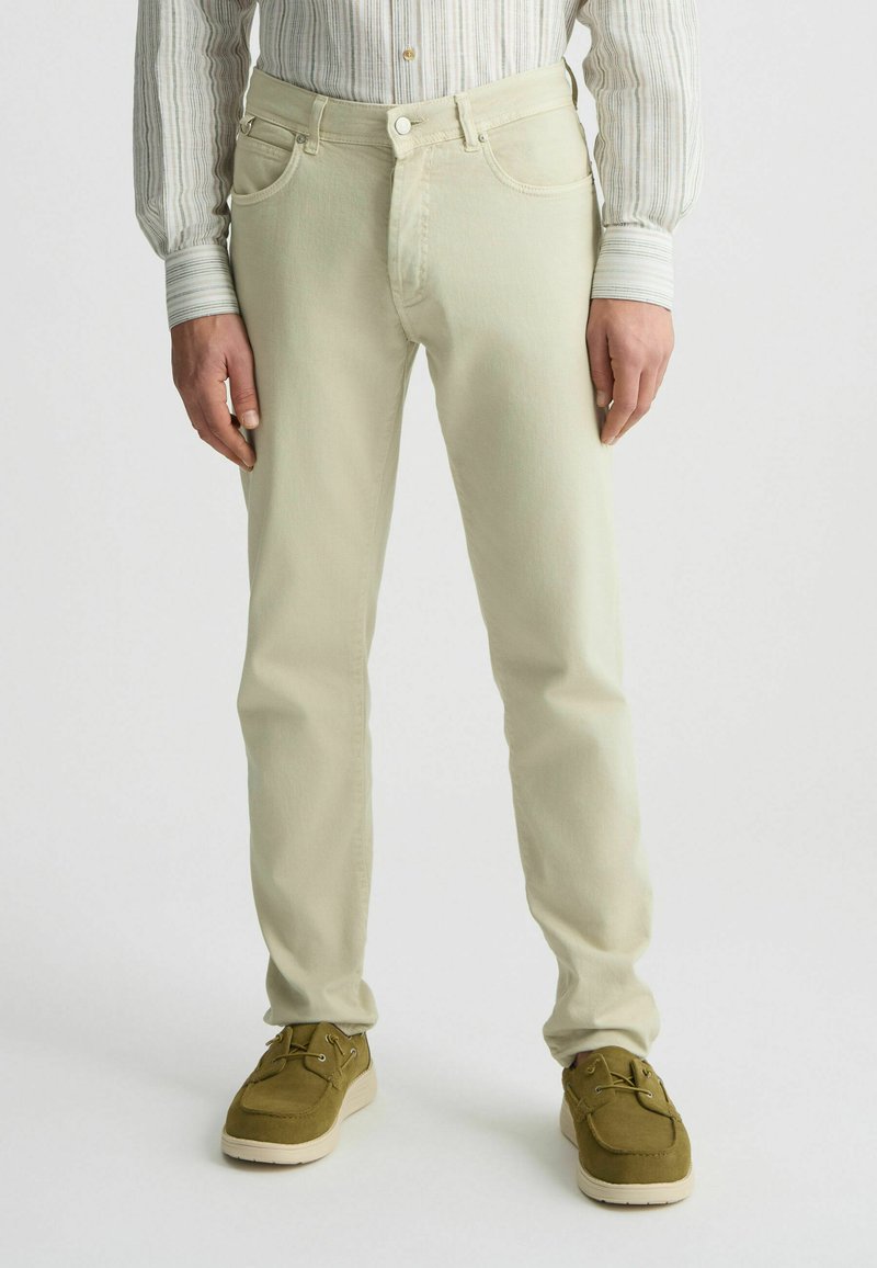 Jeans de algodón color beige claro con un corte ajustado, bolsillos sutiles y cierre de un solo botón, combinados con zapatos verde oliva con cordones.