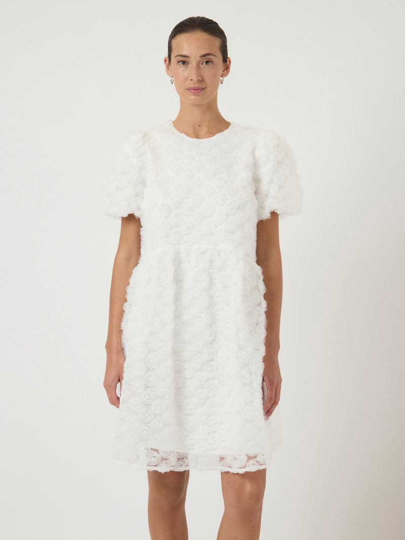 Γυναίκα ντυμένη με ένα λευκό, υφασματισμένο φόρεμα έως το γόνατο με φούσκες μανίκια και ροζ σχέδια, που στέκεται μπροστά σε έναν απλό ανοιχτό φόντο.