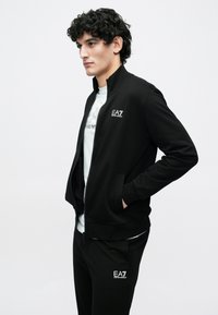 Młody mężczyzna o kręconych włosach, ubrany w czarną kurtkę na zamek i spodnie z białymi logo EA7 Emporio Armani, stoi ze spuszczonymi rękami w kieszeniach.