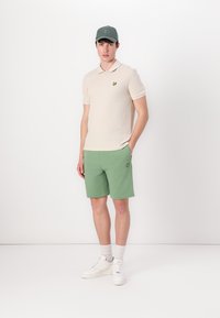 Krémové polo tričko s krátkými rukávy, zelené šortky, bílé tenisky a zelená kšiltovka. Na polo tričku i šortkách se nachází malé žluté logo.