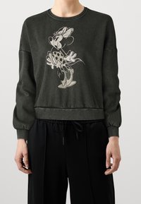 Sweat-shirt vert foncé avec manches longues bouffantes ornées d'un dessin d'un personnage tenant des fleurs. Bordures côtelées et coupe décontractée à la taille.
