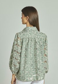 Blusa verde chiaro con motivo in pizzo floreale, maniche lunghe a palloncino e colletto. Presenta dettagli arr gathered sulla schiena e una texture leggermente trasparente.