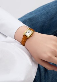 Montre rectangulaire avec un cadran blanc, des chiffres romains noirs, un boîtier doré et un bracelet en cuir brun texturé.
