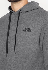 Zbliżenie szarego hoodie z czarnymi sznurkami i małym czarnym logiem "The North Face" na klatce piersiowej.