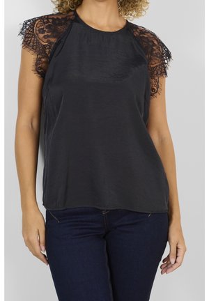 Kvinde iført en sort satinbluse med korte blonder ærmer og mørkeblå jeans mod en ensfarvet lys baggrund.