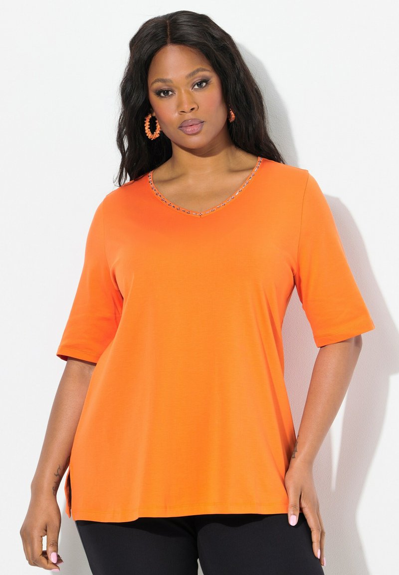 Tunic arancione a maniche corte con scollatura a V, collo decorato, spacchi laterali e una texture liscia. Indossato con pantaloni neri.