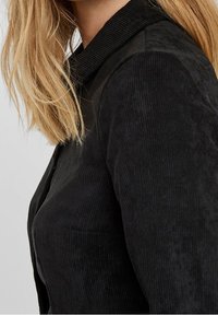 Schwarze Cordjacke mit strukturiertem Oberflächen, klassischem Kragen und knöpfbarem Vorderteil. Der Stoff wirkt weich und strapazierfähig.