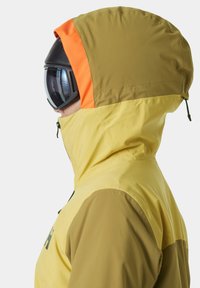 Gelbe und olivgrüne Jacke mit hohem Kragen und einer Kapuze mit orangefarbenen Akzenten, kombiniert mit schwarzen Skibrillen; glatte, technische Stofftextur.
