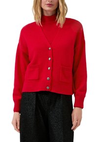Rote Strickjacke mit V-Ausschnitt, sechs silbernen Knöpfen, zwei Fronttaschen und gerippten Bündchen, kombiniert mit einem roten Rollkragenpullover und schwarzen glänzenden Hosen.
