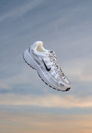 Λευκό και γκρι παπούτσι τρεξίματος Nike με το μαύρο λογότυπο Swoosh να αιωρείται πάνω σε ένα μπλε και πορτοκαλί ουρανό.