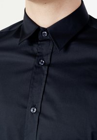 Camicetta nera con colletto classico, texture liscia e bottoni lucidi con accenti blu a contrasto.