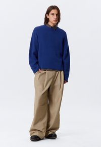 Pull en maille bleu avec des manches longues et un col côtelé, associé à un pantalon beige ample et des chaussures noires à enfiler. Design minimaliste.
