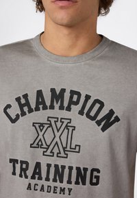 Mężczyzna w szarej koszulce z czarnym napisem: "CHAMPION XXL TRAINING ACADEMY" na piersi.