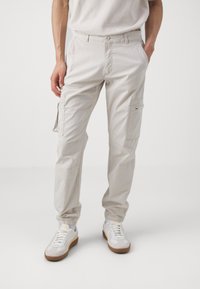 Pantalones cargo beige con puños elásticos, múltiples bolsillos laterales y un acabado texturizado. Combinados con zapatillas blancas que presentan detalles en marrón.