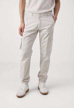 Pantalones cargo beige con puños elásticos, múltiples bolsillos laterales y un acabado texturizado. Combinados con zapatillas blancas que presentan detalles en marrón.
