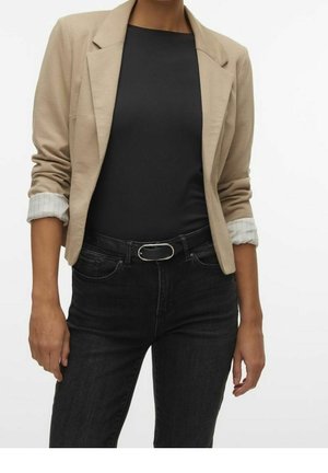 Vrouw draagt een beige blazer met opgerolde mouwen, een zwart shirt, zwarte jeans en een zwarte riem met zilveren gesp, staand tegen een witte achtergrond.