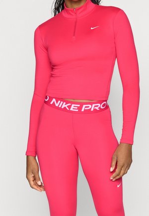 Žena nosící jasně růžový přiléhavý krátký top Nike Pro s dlouhým rukávem na zip a ladící legíny s vysokým pasem s logy Nike.