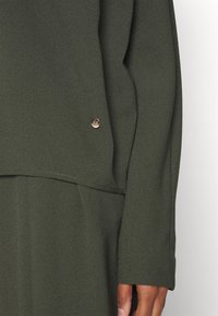 Top à manches longues vert olive foncé avec un petit logo circulaire doré près de la taille, porté avec un tissu assorti.