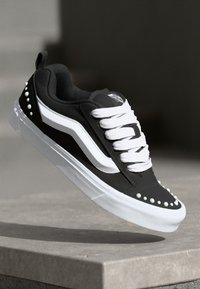 Schwarze Skate-Schuhe mit weißen Schnürsenkeln und Seitestreifen, die mit Nietenakzenten entlang der Sohle und einer Gummisohle für Grip ausgestattet sind.