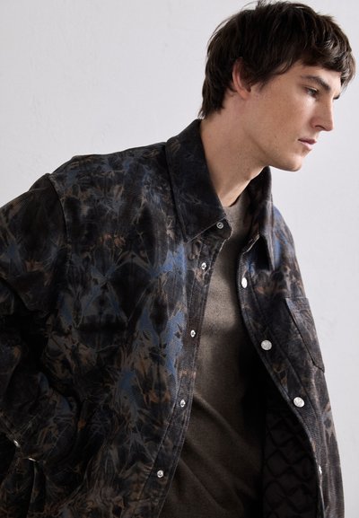 Textured, dark jacket with a floral pattern in shades of blue and brown. Features snap buttons and a quilted lining, worn over a beige shirt.