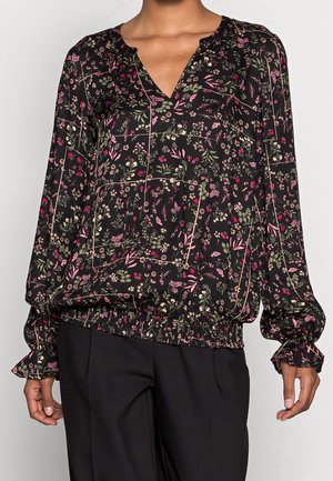 Femme portant un chemisier noir à manches longues à motifs floraux avec col en V et ourlet élastique, associé à un pantalon noir.