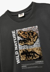 Zwarte katoenen t-shirt met een paillettenontwerp van een dinosaurus, met de tekst "WILD NATURE" in het wit ernaast gedrukt. Zorglabel zichtbaar bij de hals.