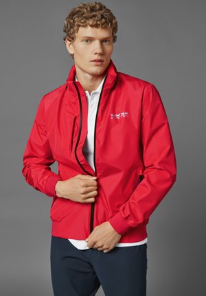 Rote leichte Jacke mit Reißverschluss, schwarzen Akzenten und Logodetails. Sie verfügt über einen Stehkragen und elastische Bündchen. Wird mit einem weißen Poloshirt getragen.