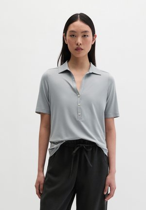 Jeune femme aux longs cheveux foncés portant un polo gris clair manches courtes et un pantalon noir avec cordon, debout devant un fond uni.