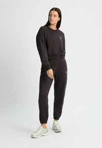 Conjunto deportivo negro que incluye un sudadera de manga larga y pantalones jogger con puños de canalé, combinado con zapatillas deportivas de color claro.