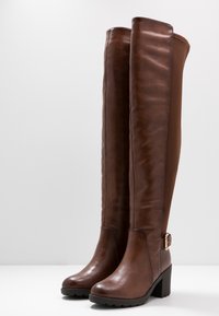 Botas marrones de cuero que llegan por encima de la rodilla, con textura suave, punta redondeada, tacón grueso y un detalle de hebilla como acento en el lateral.