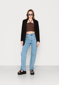 Blazer preto, top cropped castanho, jeans de cintura alta azul claro e sandálias de plataforma pretas. Os acessórios incluem óculos de sol estreitos.