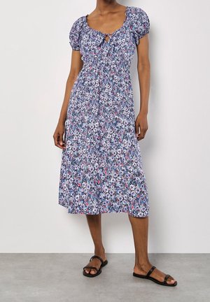 Žena v květovaných šatech ke kolenům s krátkými rukávy, se staženým pasem a černými sandály na ploché podrážce, stojící na jednobarevném pozadí.