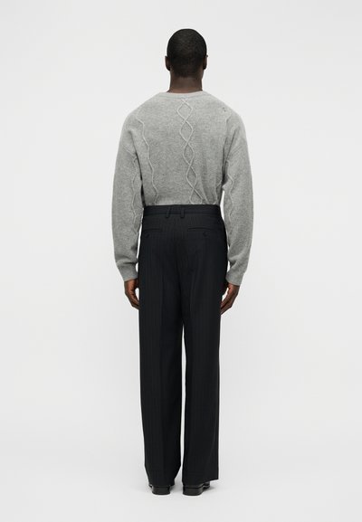 Γκρι πλεκτό πουλόβερ με σχέδιο στριμμένης πλεξούδας στην πλάτη, σε συνδυασμό με μαύρο παντελόνι με λεπτές ρίγες, που έχει άνετη εφαρμογή.