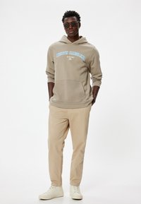 Beige hoodie med "NORTH CAROLINA" i blå tekst, kengurulomme, parret med lyse beige bukser og hvide sneakers på en ensfarvet baggrund.