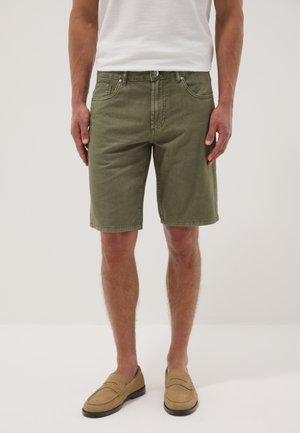 Mann trägt olivgrüne Denim-Shorts, weißes T-Shirt und beige Mokassins und steht vor einem einfarbigen hellen Hintergrund.