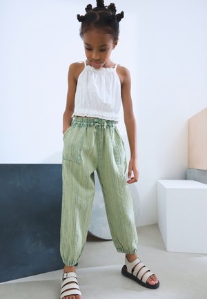 Weißes Peplum-Oberteil mit Rüschen am Ausschnitt, kombiniert mit hellgrünen gestreiften Hosen mit elastischem Bund und Seitentaschen. Weiße Sandalen.